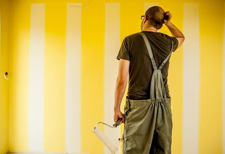 Mann in Latzhose steht mit einer Malerrolle vor gelb-weiß gestreifter Wand und ist dabei Schönheits-und Kleinreparaturen vorzunehmen.