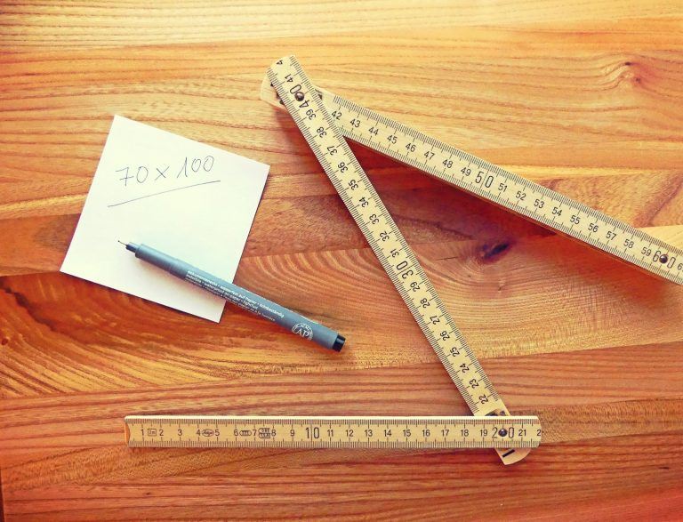 Zettel beschriftet mit Maßen, ein Stift und ein Maßstab liegen auf einem Holztisch zur Berechnung der Wohnfläche.