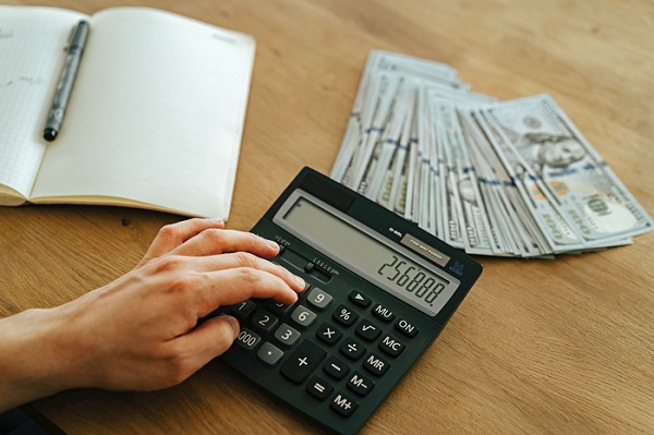 Ein Taschenrechner, eine hohe Summe Bargeld sowie ein Kalender liegen auf einem holzfarbenen Holztisch. Die Zahlen auf dem Taschenrechner stehen für ein hohes Bußgeld